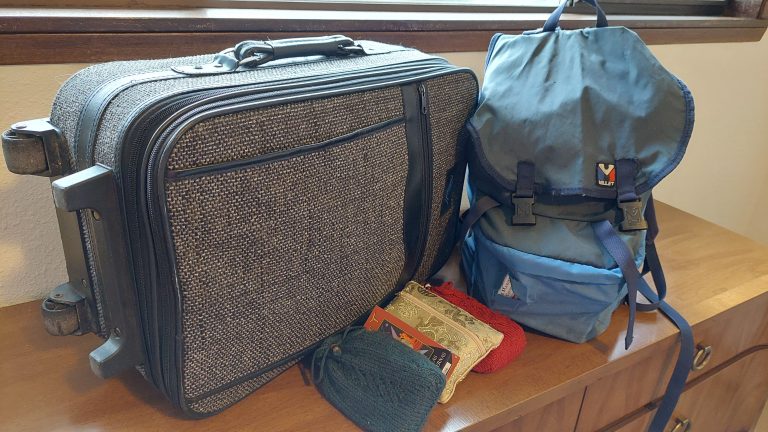 A carry-on sized suitcase and backpack with four tarot decks in front of them. The corner of another tarot deck peeks out of a hole in the front pocket of the backpack.